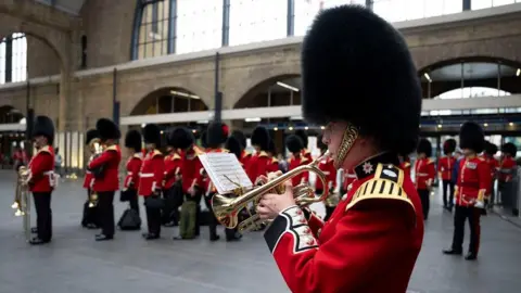 PA Media A guard in full uniform playing the trumpet in Kings Cross station. A large group of guards are standing behind him out of focus.