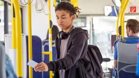 A young male on a bus holding a card to a bus ticket reader. The young man wearing a dark top and a teeshirt, with headphones around his neck.