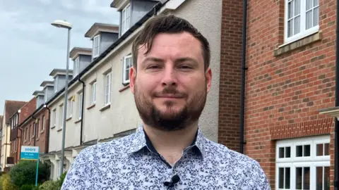 Peter Swallow, wearing a blue and white shirt and with brown hair, is stood in front of houses.