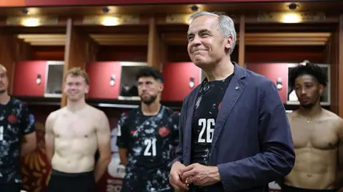 Getty Images An image showing Carney smiling in the locker room. Behind him are four players standing in front of their lockers, two of whom are shirtless and two are wearing black Canada football jerseys. Carney is wearing the same black jersey with a casual, navy blue collared sports coat on top. 