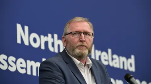 Doug Beattie is standing on a stage in front of a mic. The backdrop behind him is blue and states in white lettering: Northern Ireland Assembly. He wears glasses and has a navy suit on with a white shirt. 
