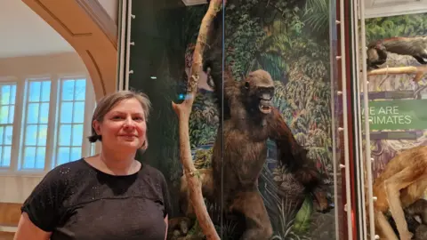 LDRS A woman standing in front of a glass museum display window behind which there is a stuffed brown gorilla grasping a tree branch
