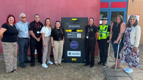 People stand either side of a new knife bin outside Doncaster Fire Station. Second from left to right: Damian Allen, CEO Doncaster Council; John Hodson, Doncaster Station Manager; Sally Jameson MP; Councillor Gemma Cobby; Clair Victory, SYFR; PC Hannah Cowling; Lisa Theobald; Kilvinder Vigurs, deputy mayor for policing and crime.
