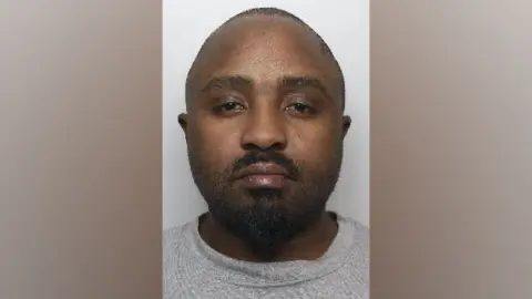 A mugshot of Pfukani, a black man who has a shaven head and a black beard. He is wearing a grey jumper