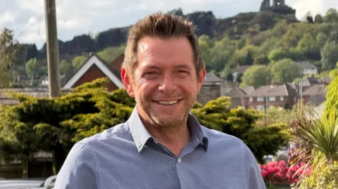 David Williams David Williams, wearing an open button shirt, smiling in front of the backdrop of his constituency, including Mow Cop Castle at the top of a hill, with houses at the bottom in the background. 