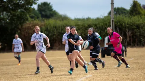 A women's rugby sevens game taking place on a pitch. A player dressed in a dark blue kit is passing the ball to her left as other players run alongside and watch her. 