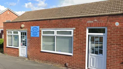 Google Doris Banham Dog Rescue's charity shop in North Hykeham. It is a small brick-built building with two front windows and a UPVC white door. 