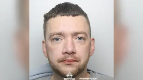 Dyfed Powys Police Custody image of Shaun Clawson. He has a short beard and brown hair which is also cut short. He is wearing a grey sweathshirt.