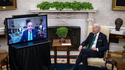 Bloomberg/Getty Images Micheál Martin pictured on a TV screen in the White House during a video call with former US President Joe Biden in March 2022.  Both men are wearing navy suits, white shirts and green ties.  A bowl of shamrock is on a table in front of Biden. 