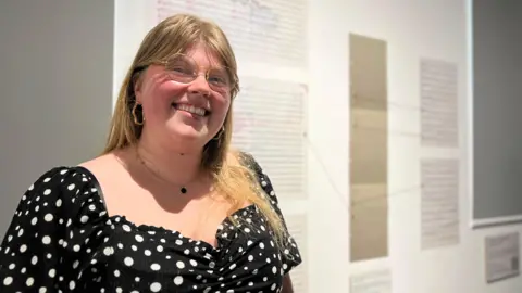 Emily smiles at the camera as she stands by some of the composition work she's done as part of the exhibition. She has a black dress on with white polka dots. She has light brown hair which comes down past her shoulders and is wearing glasses and hooped earrings.