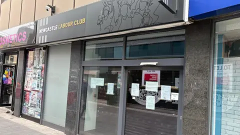 BBC The exterior of Newcastle Labour Club. The shop front has a black and white sign above the door. Shiny black lines appear to depict people standing in a crowd. There are several pieces of paper attached to the glass door. There are other shops bordering it. 