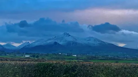 Kelly McCaughan Several mountains, their peaks topped with snow, can be seen in the distance against a cloudy sky background. In the foreground can be seen green fields and hedges.