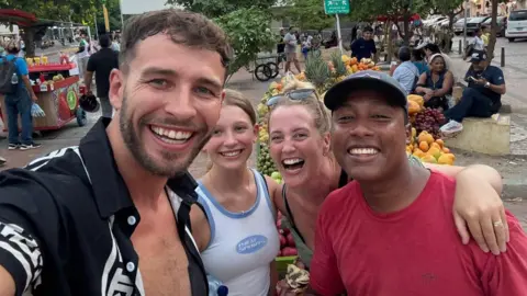 Smiths On Tour Joe, Tilly and Emma pose with another person in a selfie at an outdoor location. It appears to be a market, with large piles of fruit on a stall. A large group of people are sitting next to a road as well.