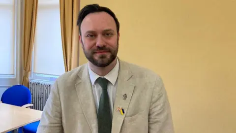 Middlesbrough councillor Luke Henman. He has short dark hair and a trimmed beard. He is wearing a cream jacket, white shirt and green tie. He has three pins on his lapel, including a union jack pin and a Ukraine flag. He is looking at the camera and smiling. He is in a room with yellow walls and yellow curtains on a window. Behind him there is a table with blue chairs.