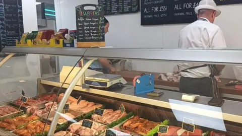 Inside a butchers shop, with glass counters displaying a variety of meat. Blackboards detail offers and prices, and two workers, wearing white uniforms and blue aprons, their backs turned, slice meat behind the display counter.