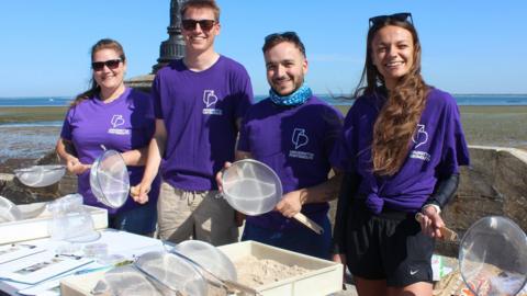 Four experts from the University of Portsmouth stood with sieves in front of the beach and with boxes of sand in front of them. They are wearing purple University of Portsmouth branded t-shirts.