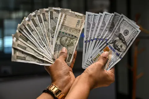 AFP via Getty Image A worker holding US dollar (R) and Indian rupee currency notes poses for a photograph at a money exchange outlet in New Delhi on April 3, 2025. US President Donald Trump ignited a potentially ruinous global trade war on April 2 as he slapped 10 percent tariffs on imports from around the world and harsh extra levies on key trading partners. (Photo by Arun SANKAR / AFP) (Photo by ARUN SANKAR/AFP via Getty Images)