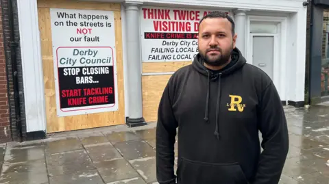 Man in black hoodie stood infront of boarded-up venue