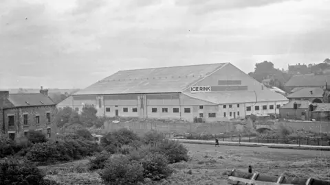 Beamish, the Living Museum of the North Black and white picture, dating from the 1940s, of Durham Ice Rink. The low rise building has a large pitched roof and long narrow windows, and a sign proclaiming Ice Rink in large letters. It is set on a river bank, with shrubs and grassland in the foreground and buildings on either side.