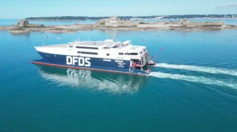 A large ferry travels through calm blue waters next to a small area of land. The ferry is large blue and white.  It has a large and white  DFDS logo printed on its side.