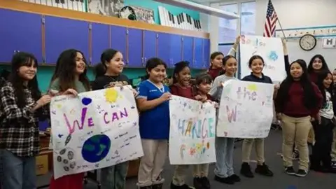 A group of children in a classroom hold handmade posters with colourful drawings and messages. They are smiling. The classroom has blue cabinets and an American flag on the wall.