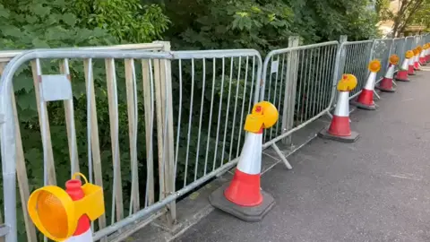 A bridge with temporary metal railings placed along the edge. Orange and white traffic cones are lined in front of the barriers. 