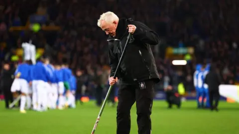 Bob Lennon, in his 60s, stands on a football pitch holding a garden hoe against the green turf. In the background a group of players wearing blue training tops are stretching, while the stands are full of spectators. Lennon has white hair and is wearing a black coat, black jeans and black shoes. 