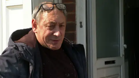 A man with grey hair and glasses on his head is standing outside a house with the door open. He is wearing a large black coat with a brown jumper