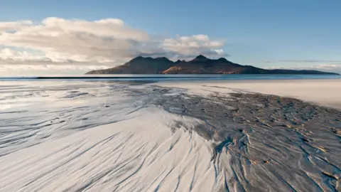 Getty Images Rum is an island of red-coloured hills and viewed across a wide sandy beach on a sunny day. There are marks in the sand left by a falling tide.
