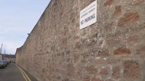 The high red sandstone boundary wall stretches down a narrow street. There is a sign on the wall that has the warning "no parking", and on the road below is a double yellow line.