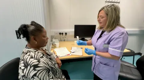 A woman sits in a doctor's surgery about to donate her saliva to a DNA database. A nurse wearing a lilac uniform stands next to her.