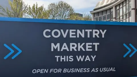 LDRS A large blue sign which reads "Coventry market - this way - open for business as usual" with some tall buildings behind it