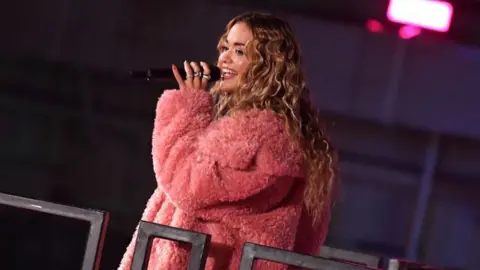 Getty Images Rita Ora performs at a VIP event celebrating the launch of her multi-season partnership with Primark wearing a pink fluffy jacket. Her hair is worn in blonde waves and she is smiling as she holds a microphone to her mouth.