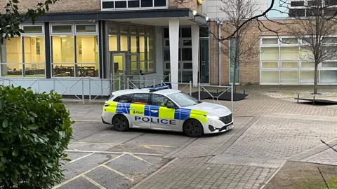 BBC/Simon Thake mobil polisi yang diparkir di halaman sekolah di luar pintu masuk gedung. Cahaya bersinar melalui ruang kelas kosong.