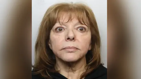 A woman with shoulder-length dark blonde hair is looking at the camera in a police custody shot. There is a Greater Manchester Police logo in the top left hand corner.