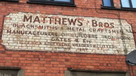 A red brick wall with brown framed windows below and above and in between a sign painted on it. The sign is a cream rectangle and painted on it says "Matthews Bros, blacksmiths & metal craftsmen, manufacturers, builders, ironworks."