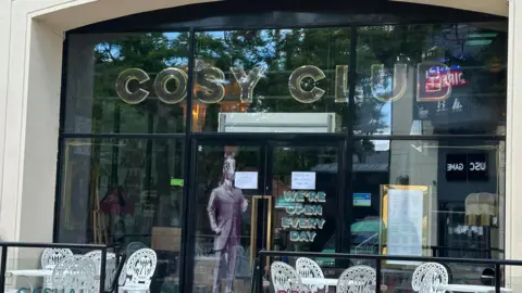 The front of the Cosy Club bar. There is a sign saying Cosy Club. Hand written posters have been put up in the window saying "sorry we are closed".