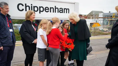 Ben Birchall/PA Wire Queen Camilla wearing a green dress and holding a black bag shakes hands with a young girl holding a poppy wreath. A crowd of people surround them and a railway sign reads "Chippenham".
