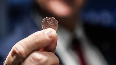 A man holds up a penny between his thumb and fore finger very close to the camera.