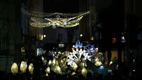 Getty Images Dozens of people holding up lanterns. There are buildings either side of the street. 