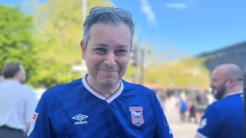 Aimee Dexter/BBC Robin Collins is standing to the left. He has short grey hair and has sunglasses resting on top of his head. He is wearing a blue and white Ipswich Town T-shirt.