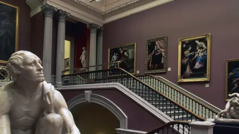 Getty Images A number of portraits in gold frames adorn the Staircase Hall in Dublin's National Gallery. A statue of a man crouched down looking to the sky is in the foreground and another statue of two men wrestling is at the bottom of the staircase. The top of the staircase has four large pillars and a statue of a man standing upright.