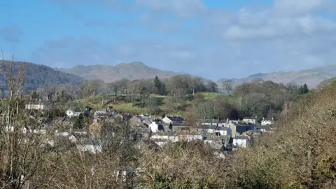 sammyadd The rooftops of homes in Broughton in Furness pictured on a sunny day, with blue skies. In the background there are trees, green fields and the fells rising off into the distance.