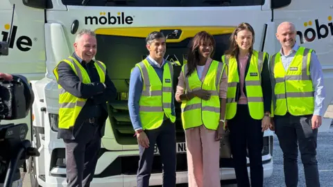 A group of five people, two women and three men, stand in front of a lorry. They all wear green high-vis jackets.