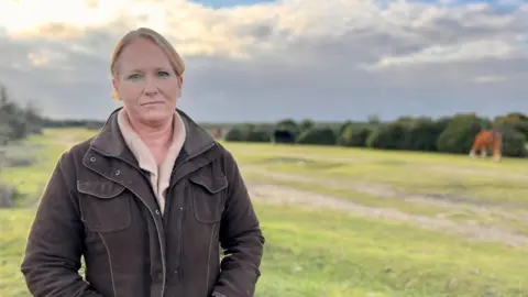 Sally Jolly, a middle-aged woman standing in the forest. She is wearing a beige jumper and brown coat and is standing with her hands in her pockets. Her blonde hair is tied back in a ponytail. The forest behind her is out of focus, but a brown pony can be seen grazing on some grass.