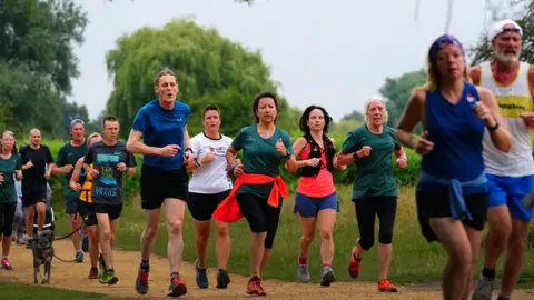 Runners are pictured in a park following each other on a gravel path. Many wear T-shirts and shorts. Greenery and trees can be seen behind them.