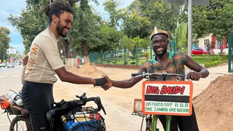 Contributed A man sitting on a bike shakes hands with another man walking alongside a bike. The man on the left has black dreadlocks that have been tied back and he wears a cream cycling jersey, black cycling shorts and black gloves. The man on the right wears a hat and a sleeveless chequered shirt. There is a sign on the front of his bike with a mobile number and the name of Big Baudi. 