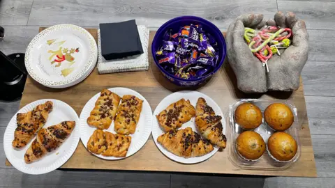 Image shows a variety of snacks. On the bottom left are chocolate filled croissants, then to the right of them is an apple turnover, and to the right of that more chocolate filled croissants and an apple turnover, then to the right of those are a box of 4 chocolate chip muffins. On the top right is an ornament of two hands which contain candy canes and to the left of those are a tub of Heroes. There are napkins to the left of those and plates to the left of those. 