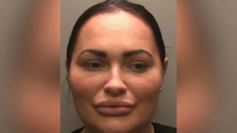 A police custody picture of Natasha Allarakhia. She has black hair tied back and is looking to the side. The wall behind her is grey. 