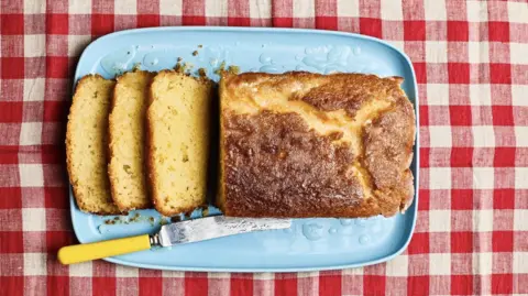 Mowie Kay A yellow loaf cake on a blue plate with three slices cut off the end and a yellow knife laying beside it and the plate is on a red and white checked tablecloth
